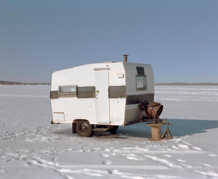 A white ice fishing shack sits on a vast expanse of snow and ice under a clear sky.