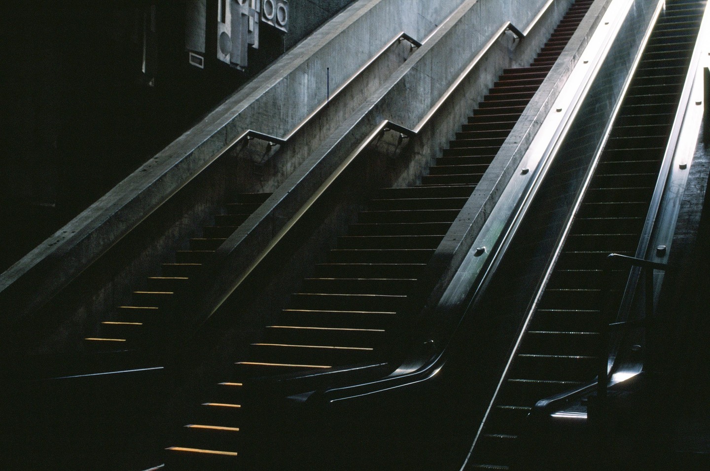 Sunlight illuminates the steps of a dark concrete staircase and escalator.