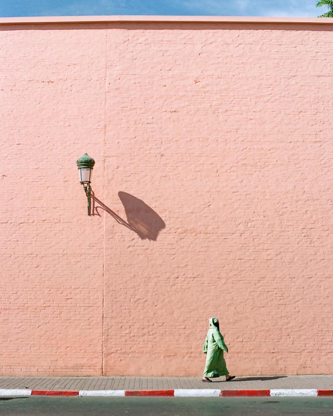 A person in a green robe walks along a sidewalk next to a pink wall with a street lamp casting a shadow.