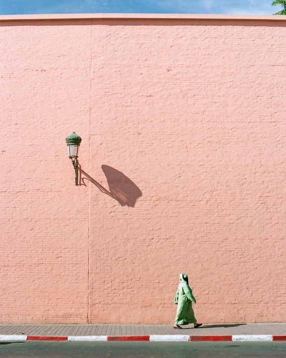 A person in a green robe walks along a sidewalk next to a pink wall with a street lamp casting a shadow.