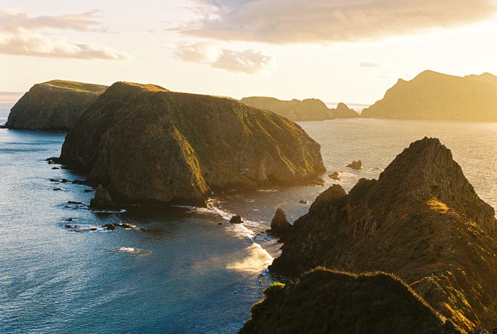 A golden hour landscape shows several rocky islands and the ocean under a bright sky.