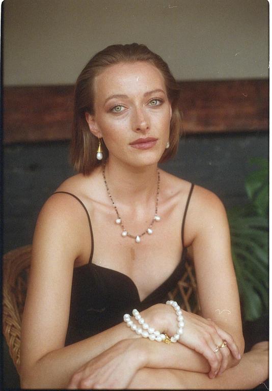 A woman wearing a black dress and pearl jewelry looks directly at the viewer.