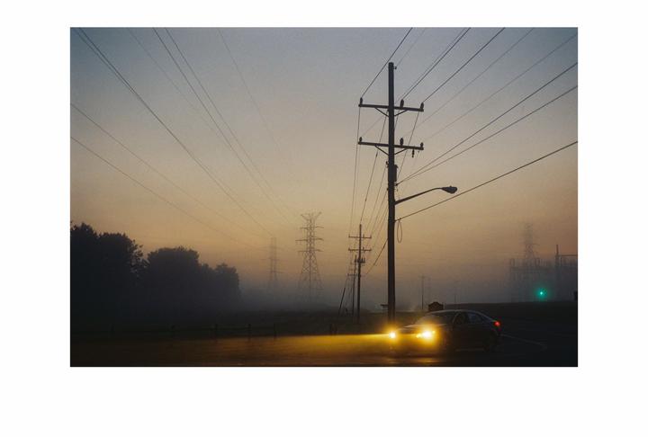 A car with its headlights on drives on a road with power lines and transmission towers in the fog at dawn.