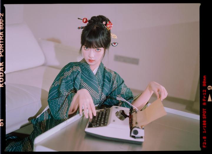 A woman in a green kimono with hair ornaments sits at a table with a typewriter.