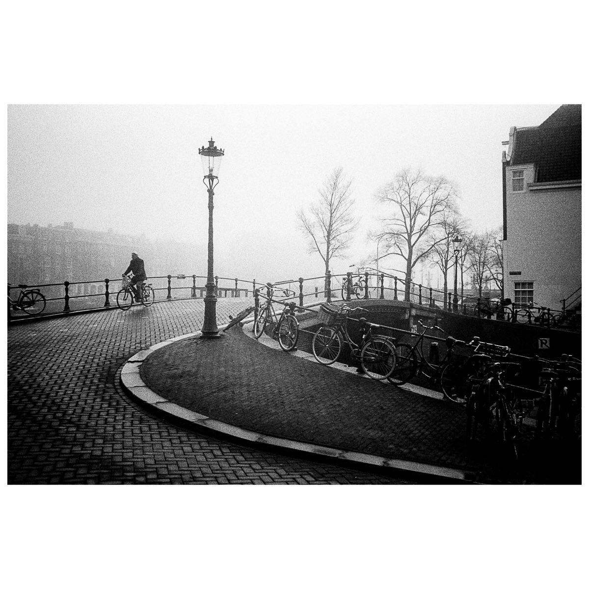 A man rides a bicycle across a cobblestone bridge in a hazy, black and white street scene with many parked bicycles and a streetlamp.