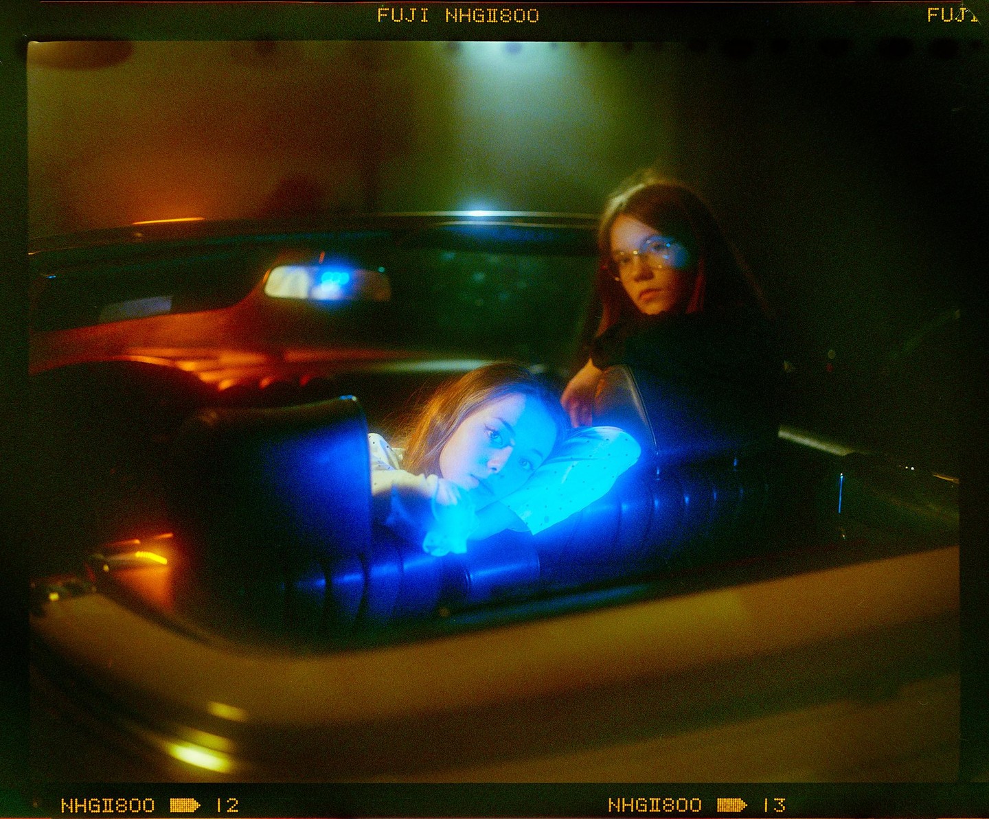 Two girls are sitting in a car at night, one of them illuminated by blue light.
