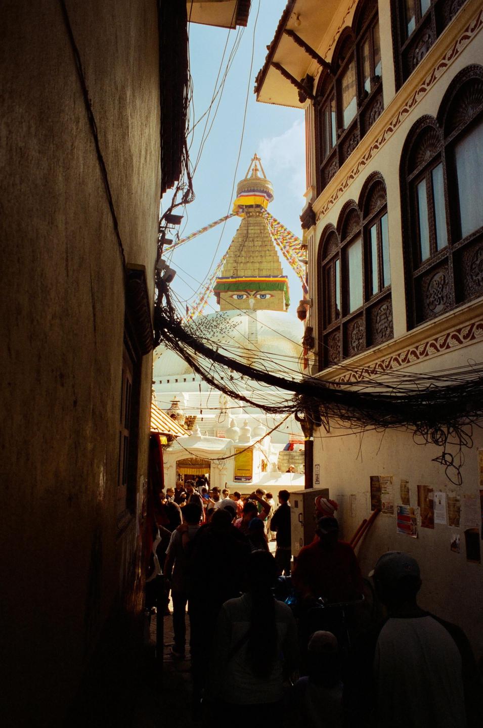 A narrow alleyway with many people leads to a large white stupa with a golden spire and painted eyes.