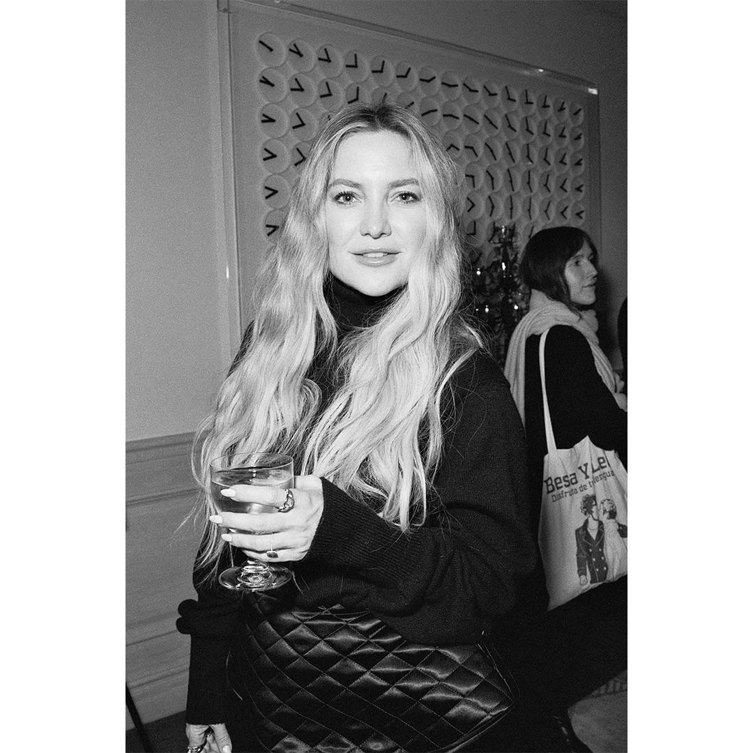 A woman with long wavy hair holds a glass and smiles at the camera.