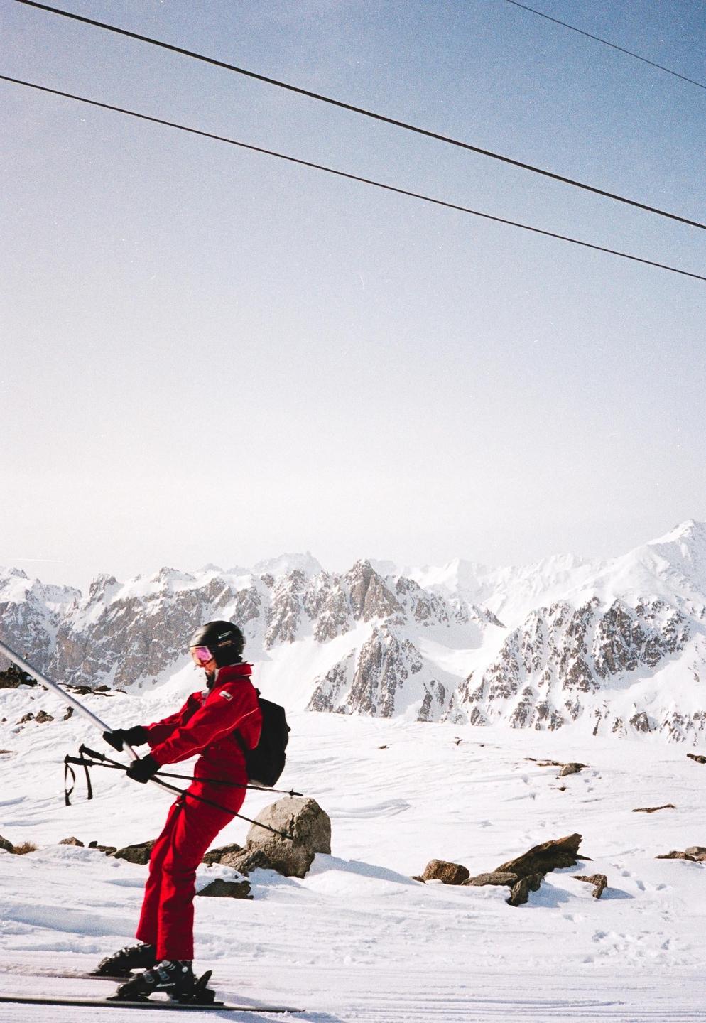 A skier in a red suit is riding a ski lift with snow-covered mountains in the background.
