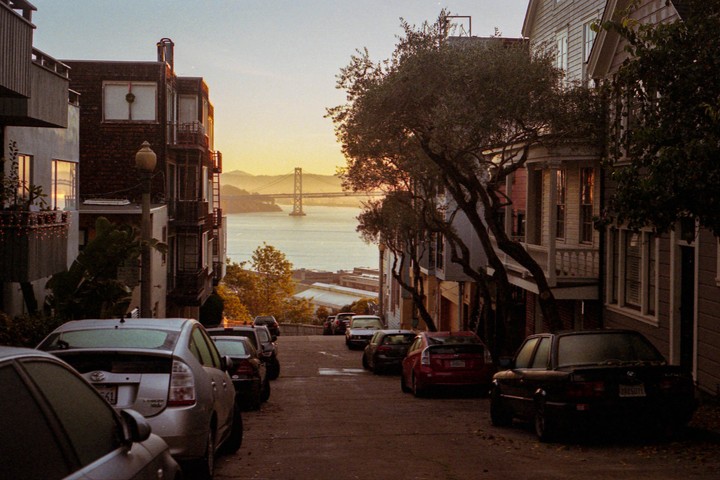 A street lined with parked cars and buildings leads down to a body of water with a bridge in the distance at sunrise.