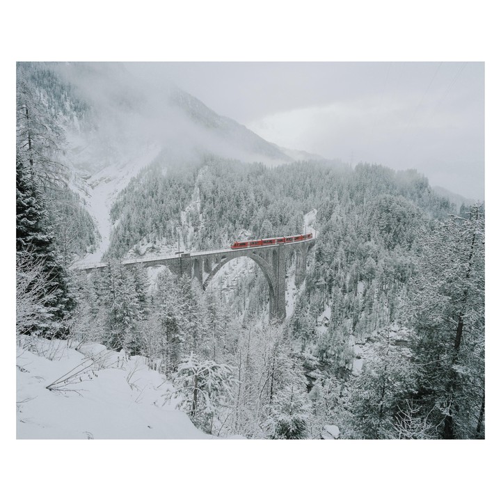 A red train crosses a tall stone bridge over a snowy valley filled with pine trees and fog.
