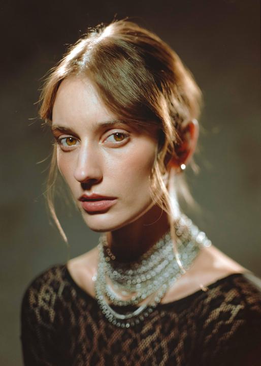 A woman with light brown hair and a multi-strand necklace looks directly at the viewer.
