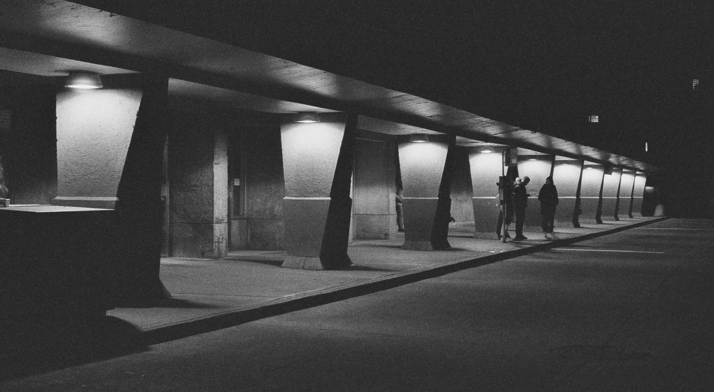 Three people stand under a concrete structure with a row of lights illuminating the pillars at night.