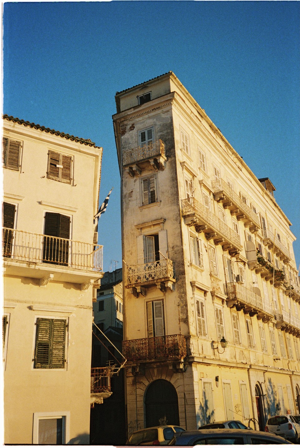 Two buildings with balconies and shutters are visible under a clear blue sky, with a Greek flag hanging between them.