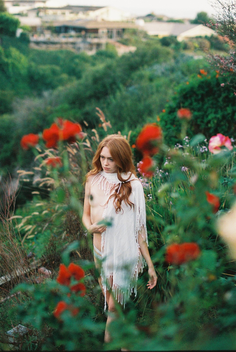A woman with long red hair and a white shawl stands in a garden with red flowers.