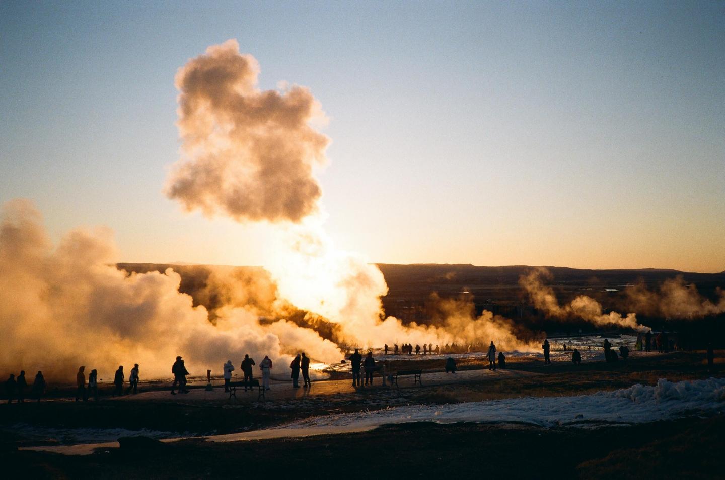 A crowd of people watches a geyser erupting at sunset, with steam and golden light filling the air.