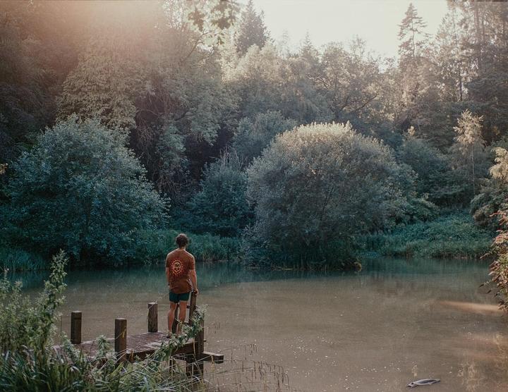 A man stands on a wooden dock overlooking a lake surrounded by trees.