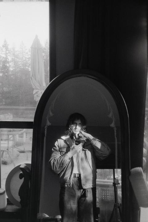 A man with long hair wearing a denim jacket takes a self-portrait in a mirror.