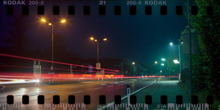 A long exposure photograph captures light trails from vehicles on a street at night, illuminated by streetlights.