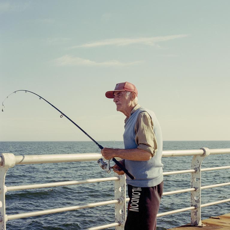 A man in a red cap and blue vest stands on a pier holding a fishing rod over the ocean.