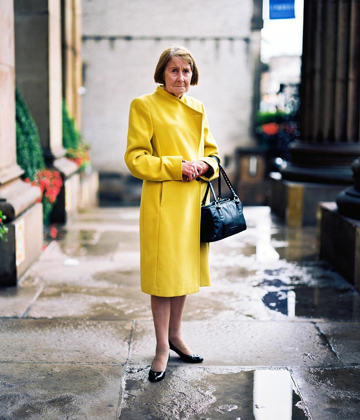 A woman in a yellow coat stands on a wet street holding a handbag.