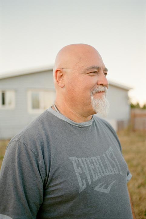 A man with a white beard and a gray t-shirt stands outdoors, looking to the right.