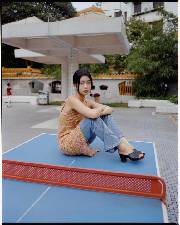 A woman sits on a blue ping-pong table with a red net, looking at the viewer.