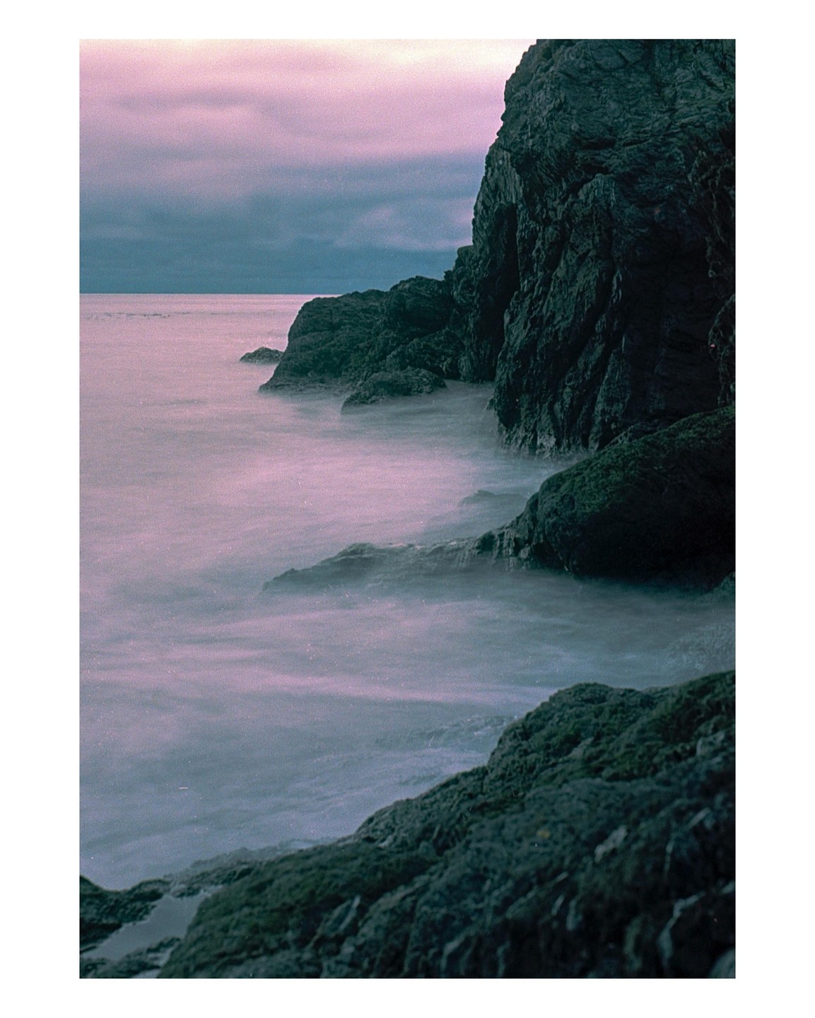 A long exposure shot captures the movement of water around dark, rocky cliffs under a purple sky.