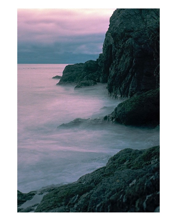 A long exposure shot captures the movement of water around dark, rocky cliffs under a purple sky.