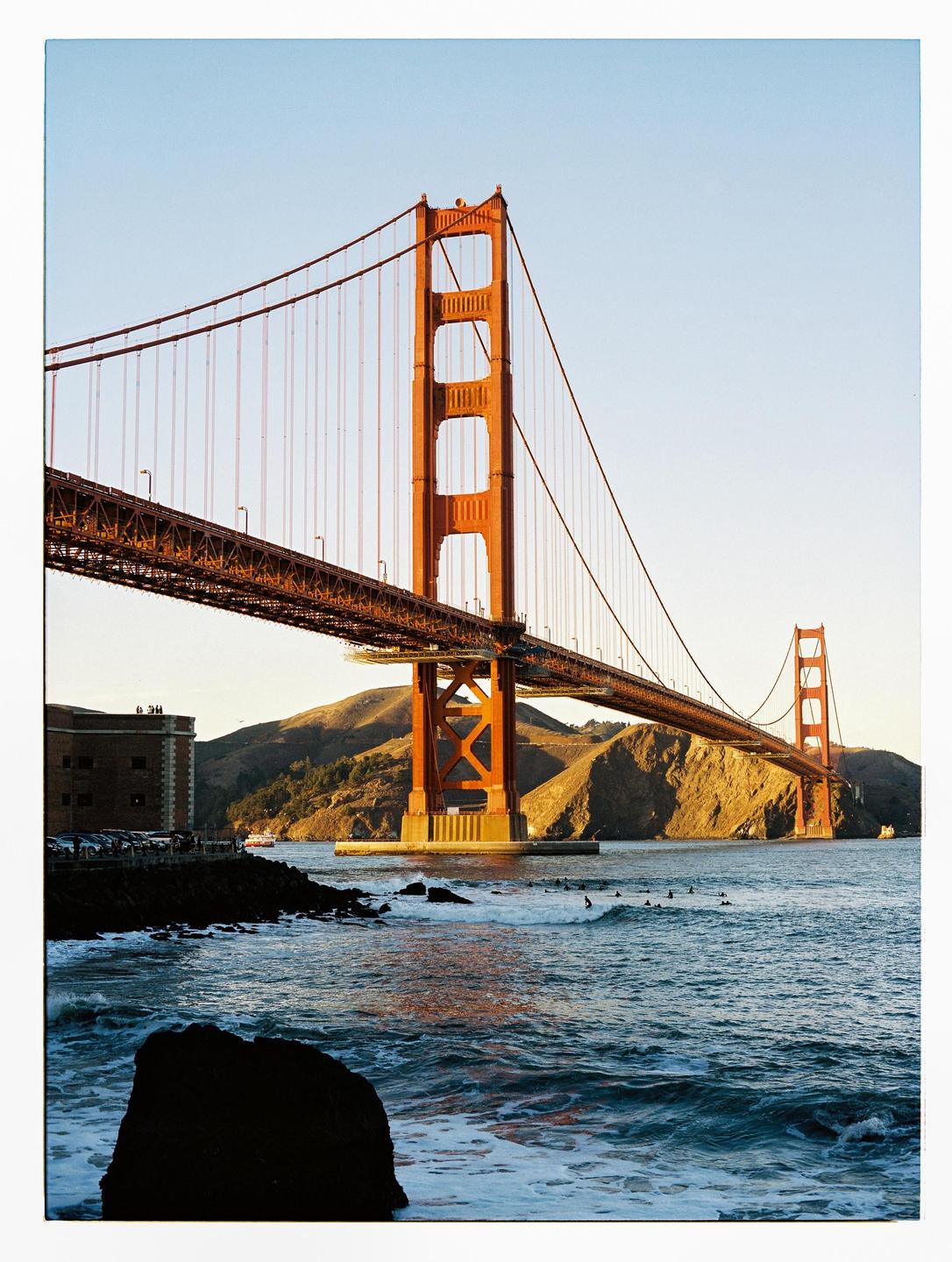 The Golden Gate Bridge spans a body of water with surfers and mountains in the background during golden hour.