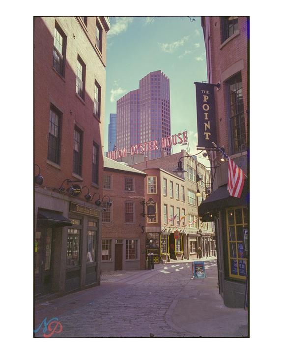 A narrow cobblestone street is lined with brick buildings and shops, with a tall modern building visible in the background.