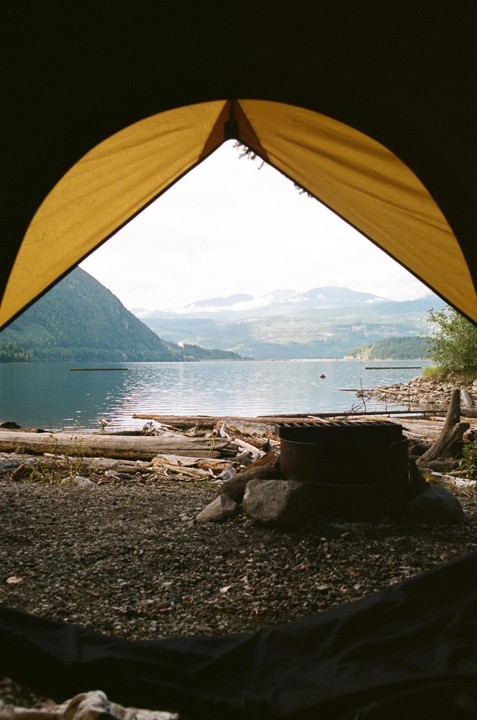 A view from inside a tent shows a lake, mountains, and a fire pit on a rocky shore.