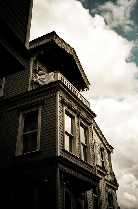 A woman in a white dress leans over a balcony railing on a wooden house under a cloudy sky.