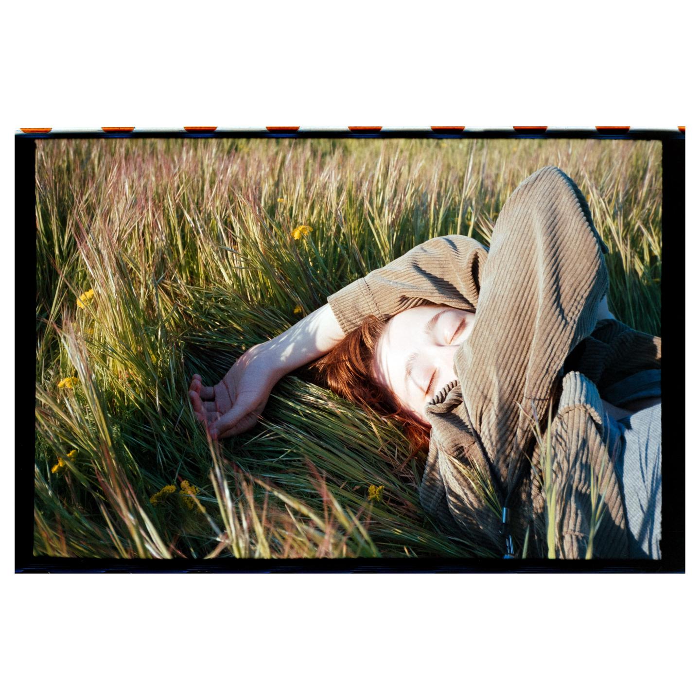 A woman with red hair is lying in tall grass with her eyes closed and one arm covering her face.