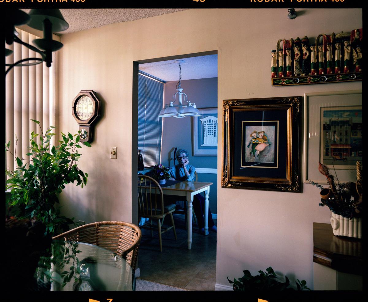 A woman sits at a dining table in a room with a wall clock and framed artwork.