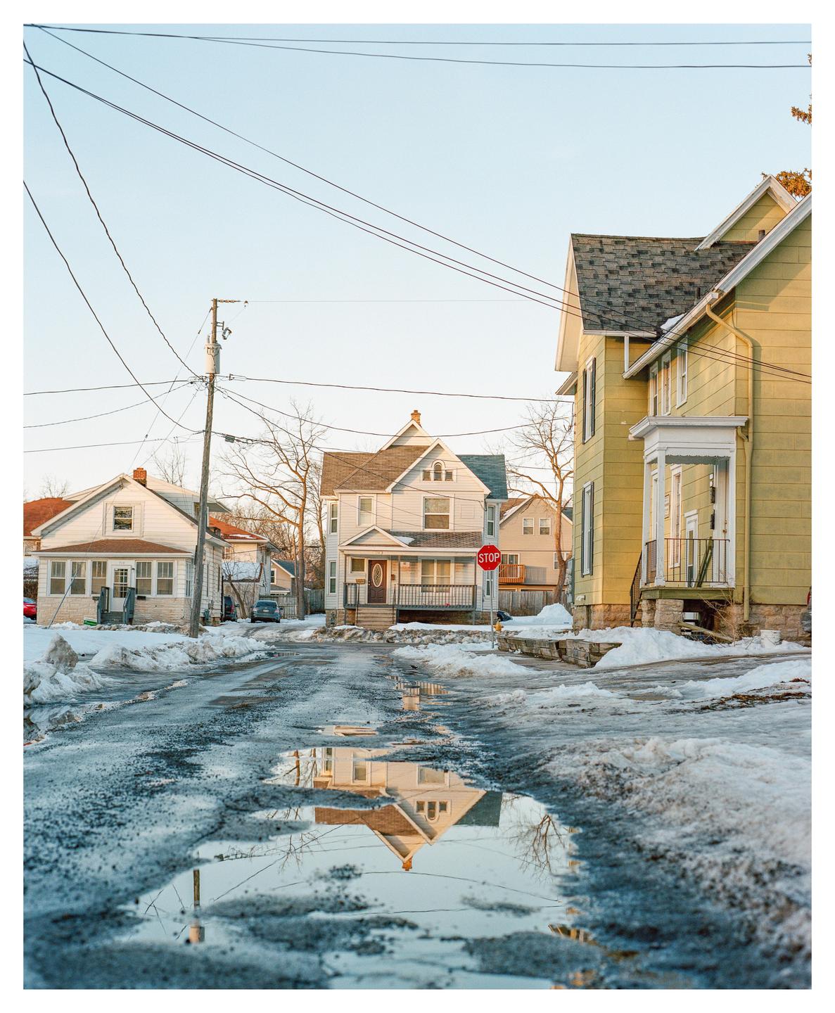 A street with melting snow and puddles reflects houses under a clear sky.