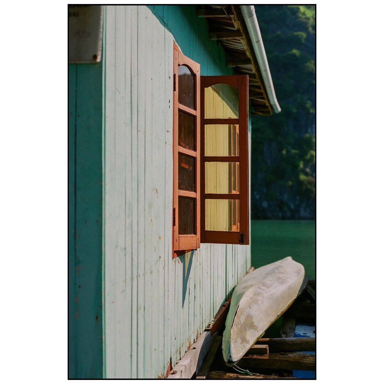A wooden house with an open window and a boat resting against its side is shown on the water.