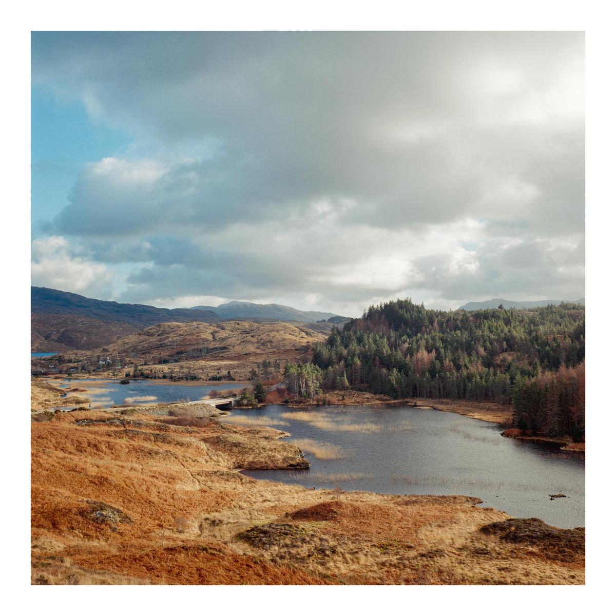 A body of water is surrounded by hills and a forest under a cloudy sky.