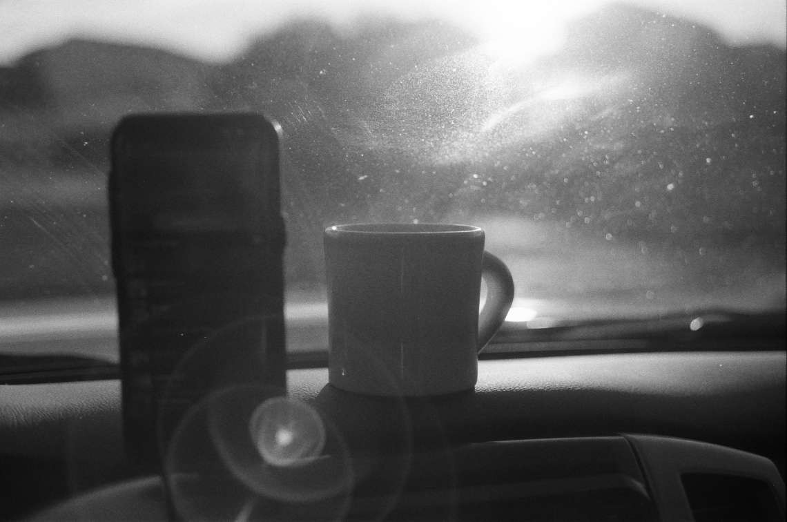 A mug and a phone are on a car dashboard, with light flaring through the windshield.