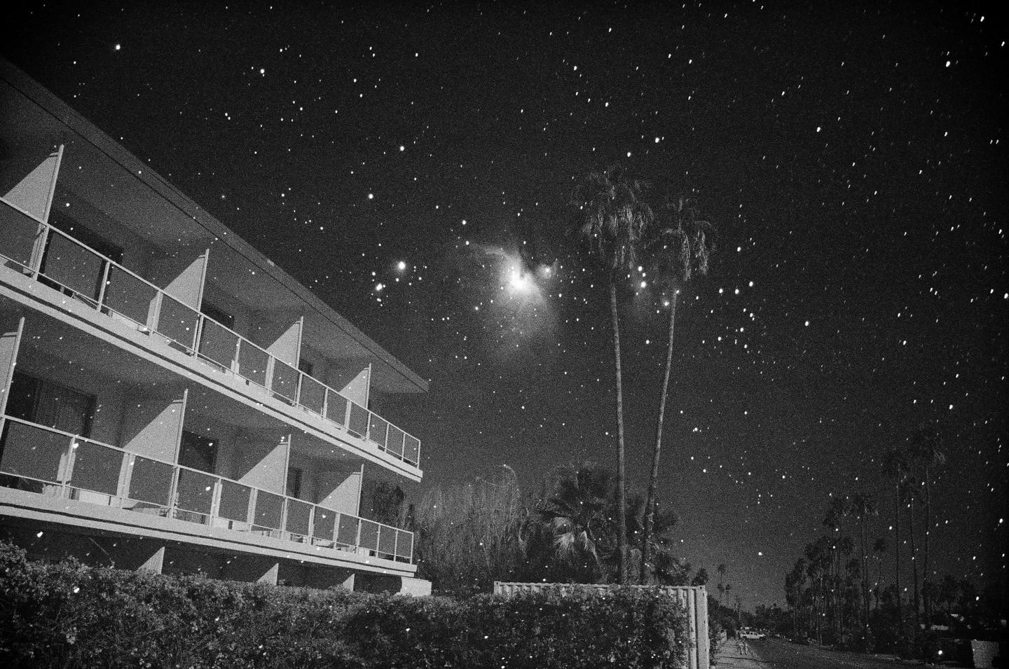 A multi-story building with balconies stands under a starry night sky with palm trees and a street visible.