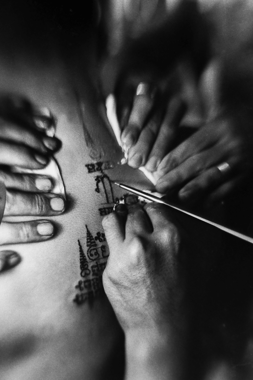 A person's torso is being tattooed with a traditional design by two sets of hands, one holding the tattooing tool and the other stretching the skin.