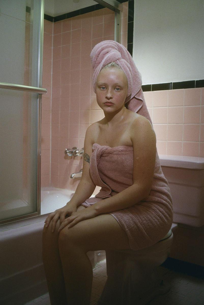 A woman wrapped in a pink towel sits on a toilet in a pink tiled bathroom.