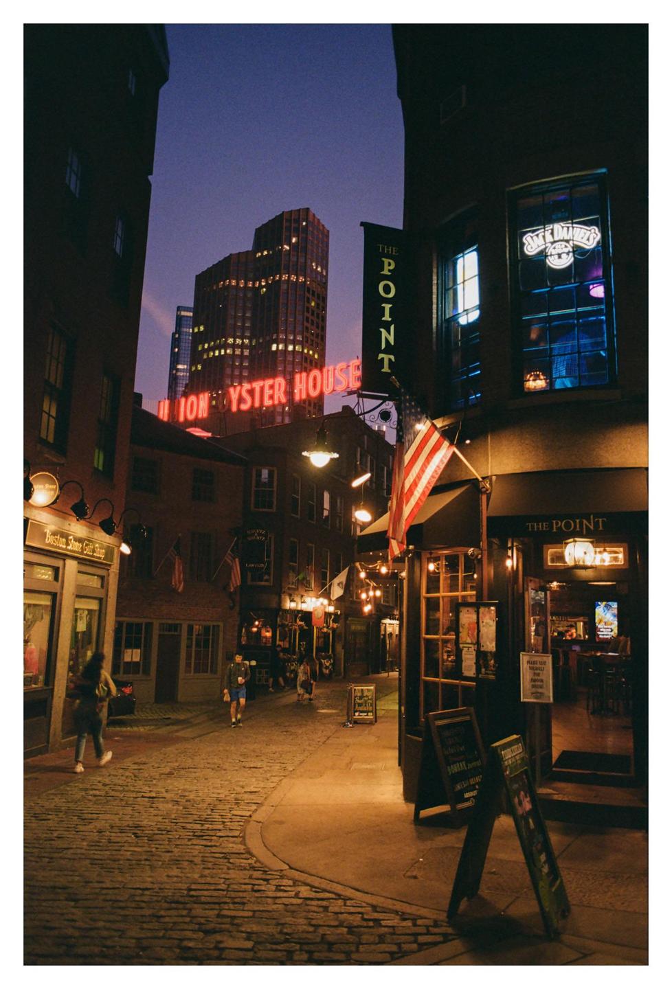 A cobblestone street is illuminated by neon signs and string lights at night, with people walking along the street.