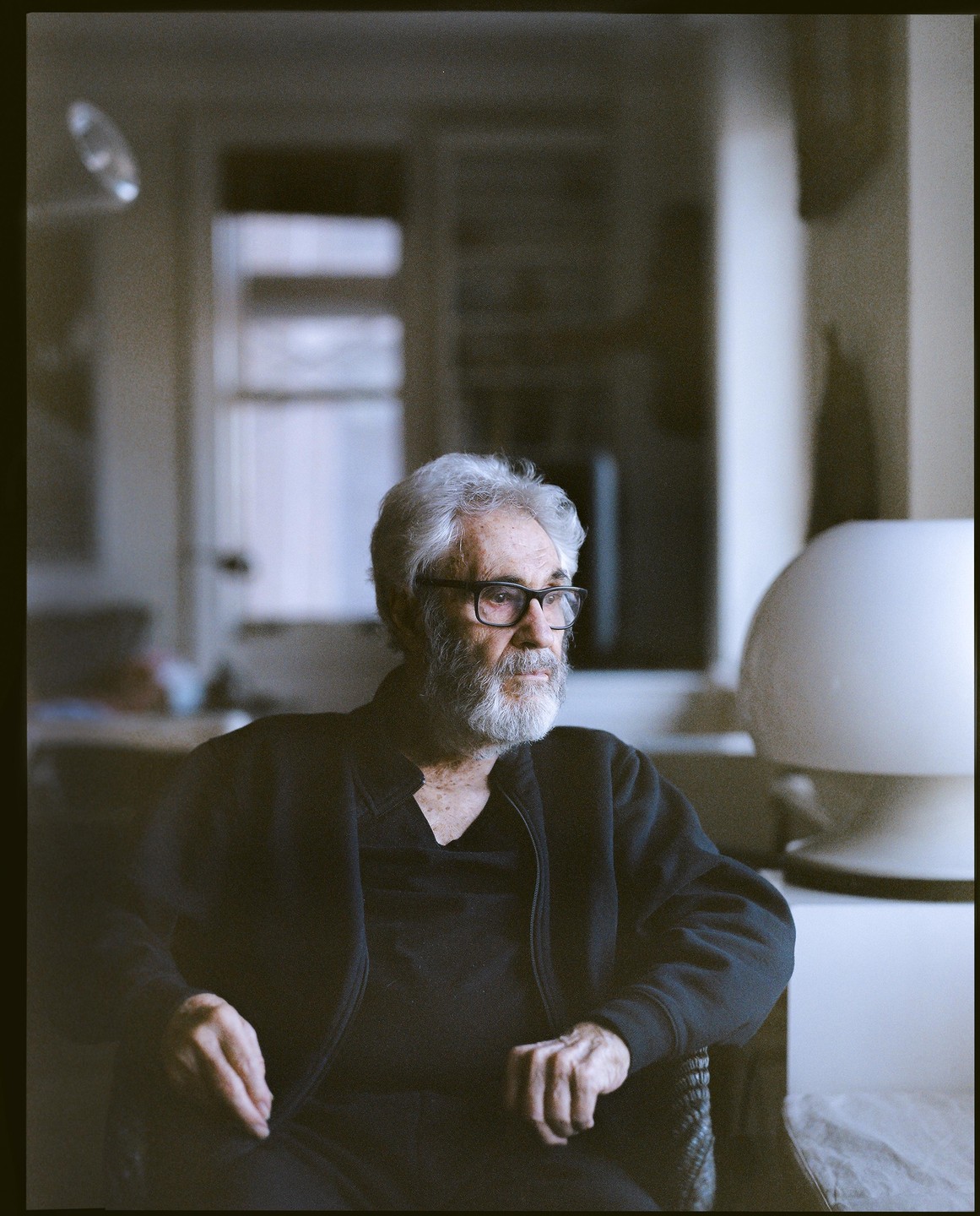 A man with a white beard and glasses sits in a chair looking to the right.