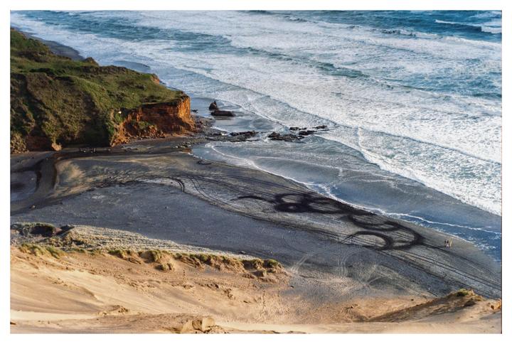 An aerial view shows a beach with tire tracks on the sand, a cliff, and ocean waves.