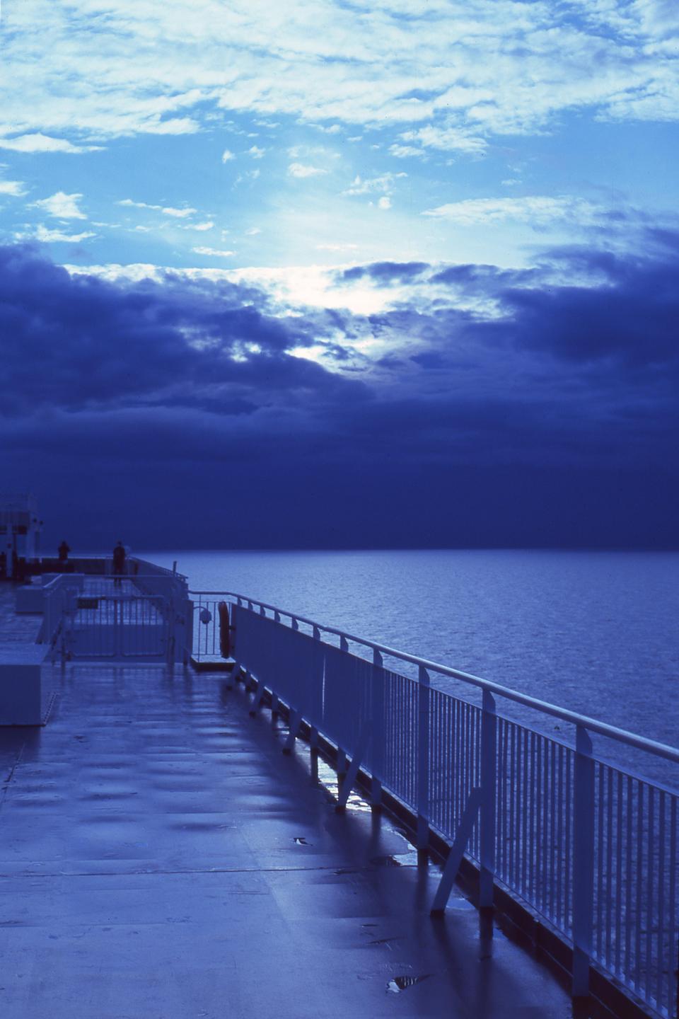 A ferry deck with a railing overlooks a vast expanse of ocean under a cloudy sky.