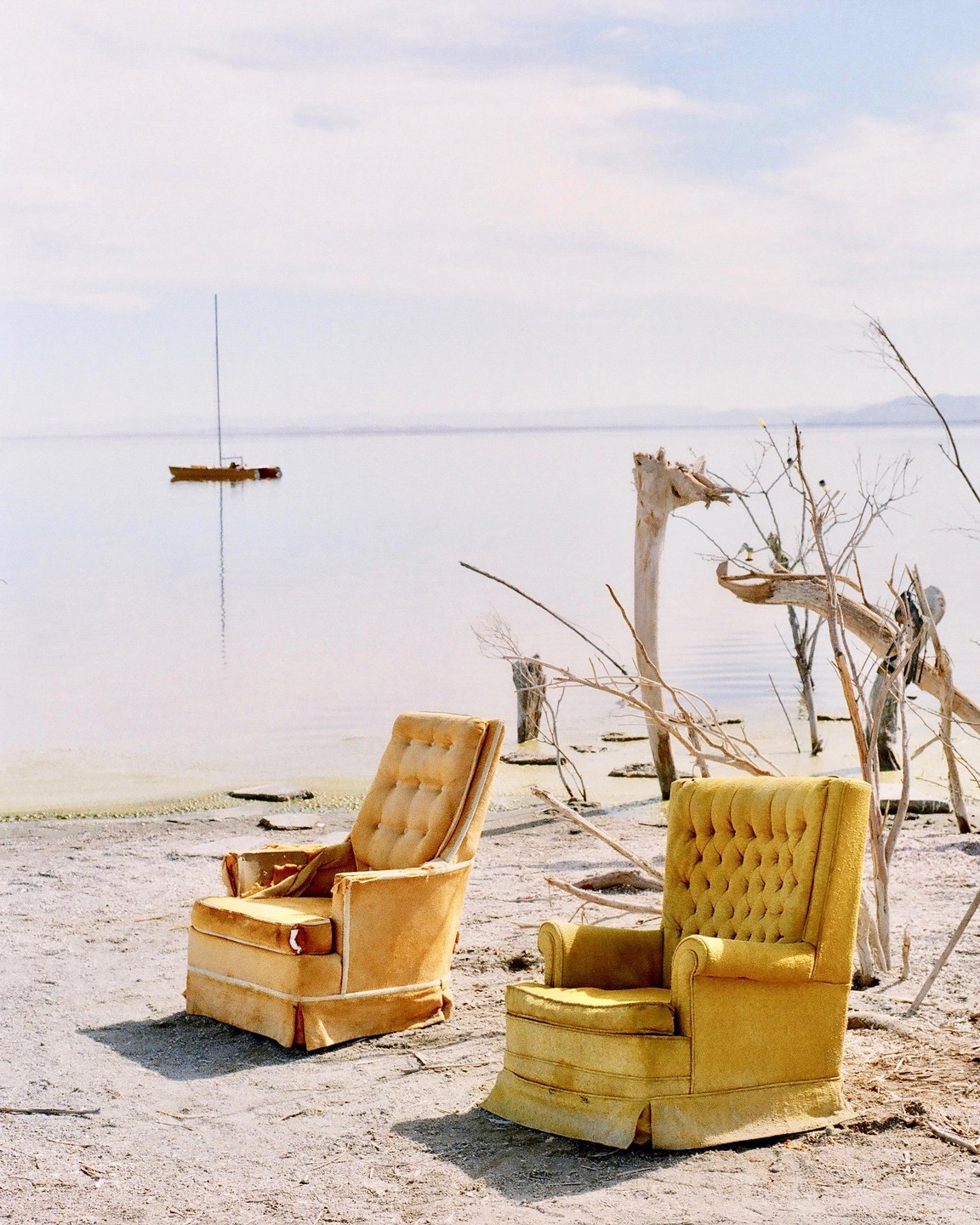 Two yellow armchairs sit on a sandy beach next to a body of water with a sailboat in the distance.