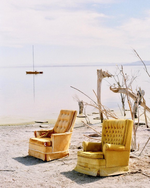 Two yellow armchairs sit on a sandy beach next to a body of water with a sailboat in the distance.
