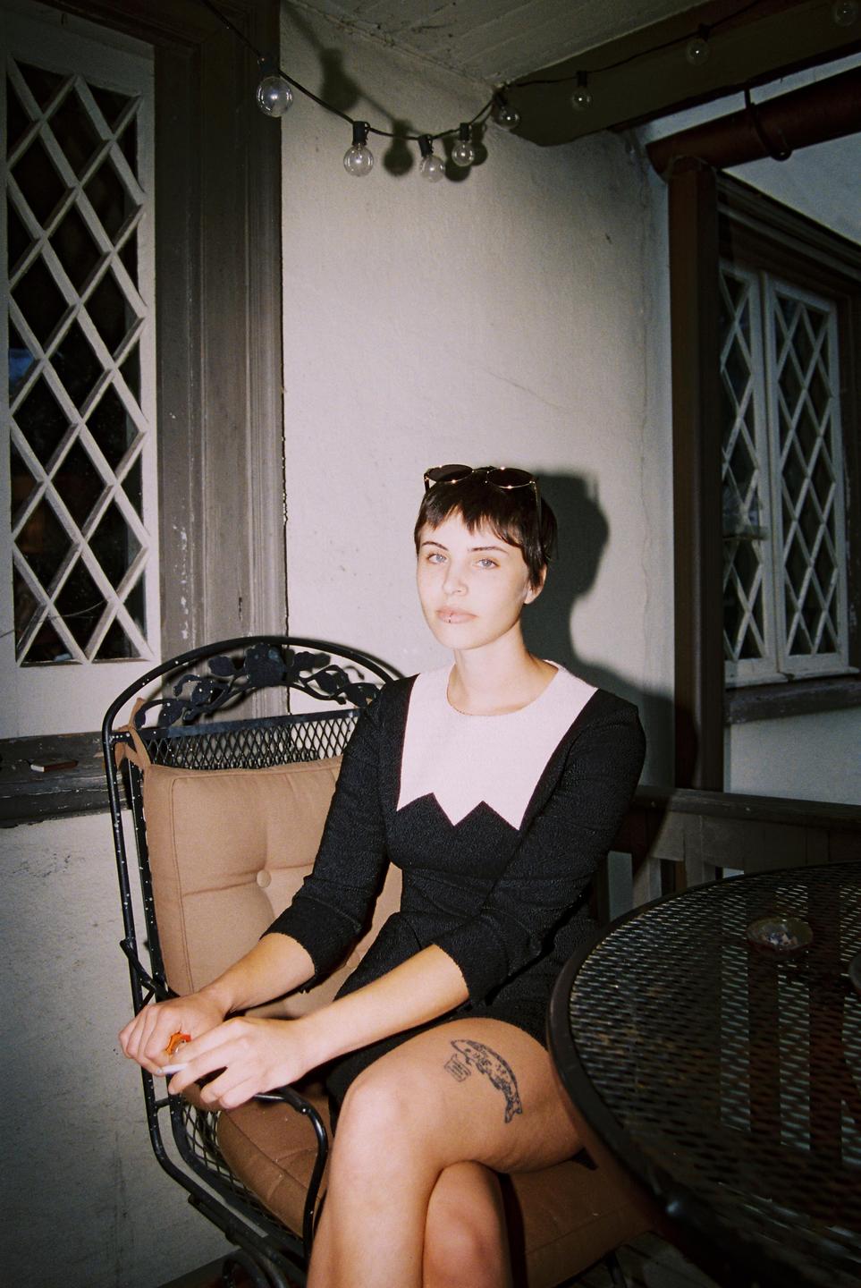 A woman with short hair and sunglasses on her head sits on a patio chair next to a table.