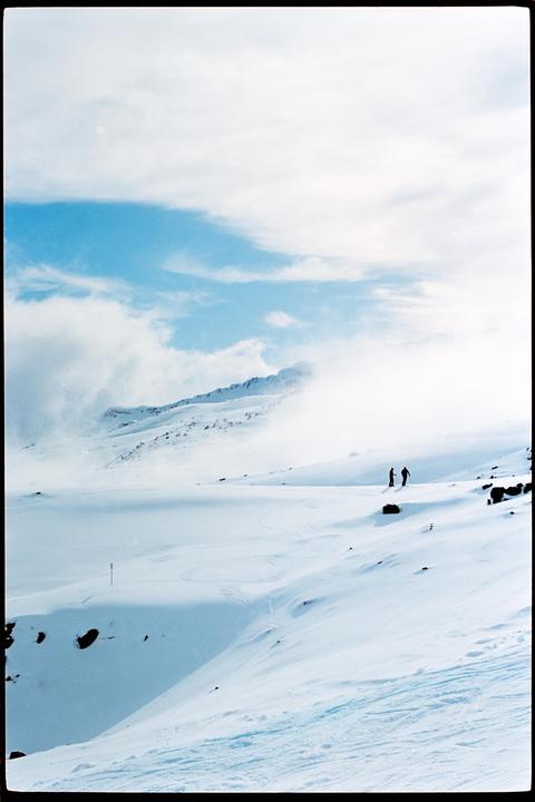 Two people stand on a snow-covered mountain with clouds in the background.